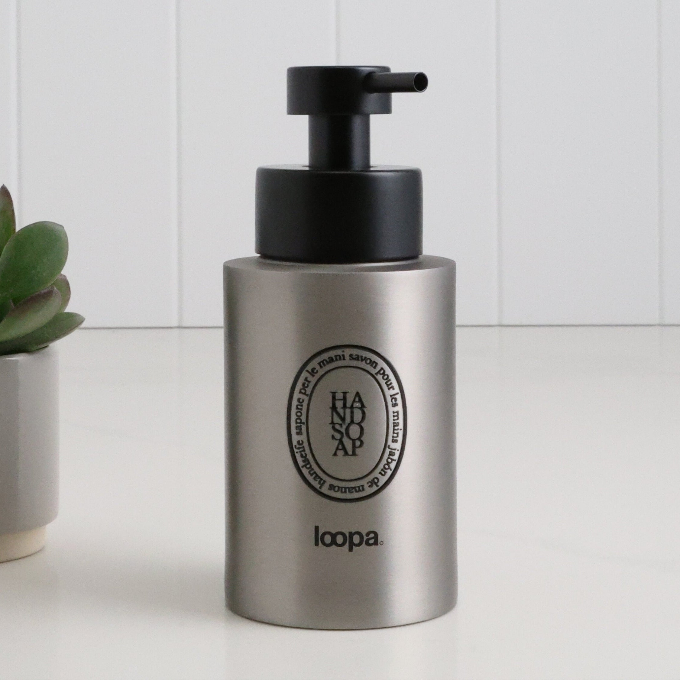 Brushed stainless steel round bottle dispenser with a black oval label, shown as a 250ml refillable bottle suited to cloakrooms, WC areas, or compact bathrooms