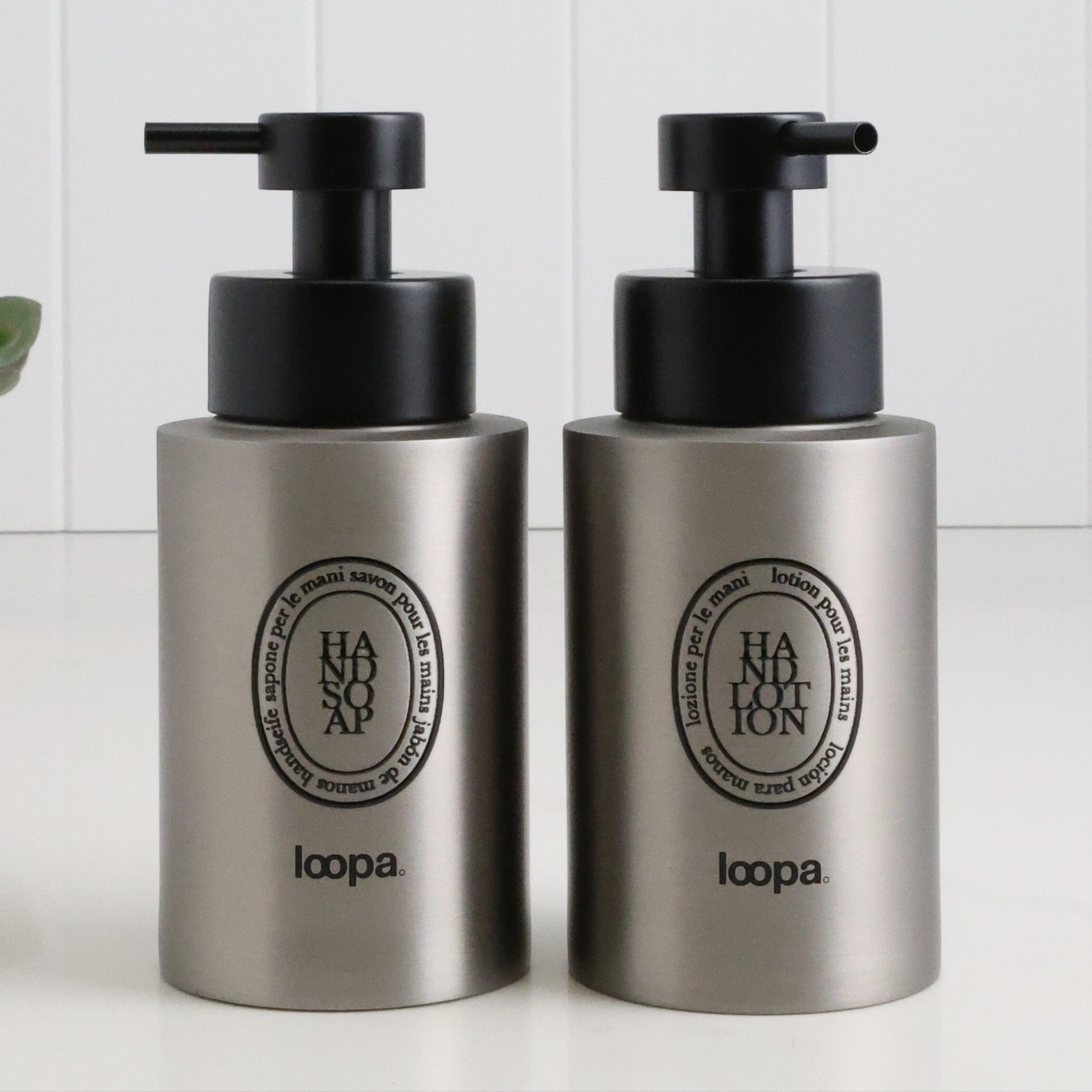 Two brushed stainless steel round bottle dispensers with black oval labels, shown as matching 250ml refillable bottles for bathrooms, cloakrooms, or WC spaces