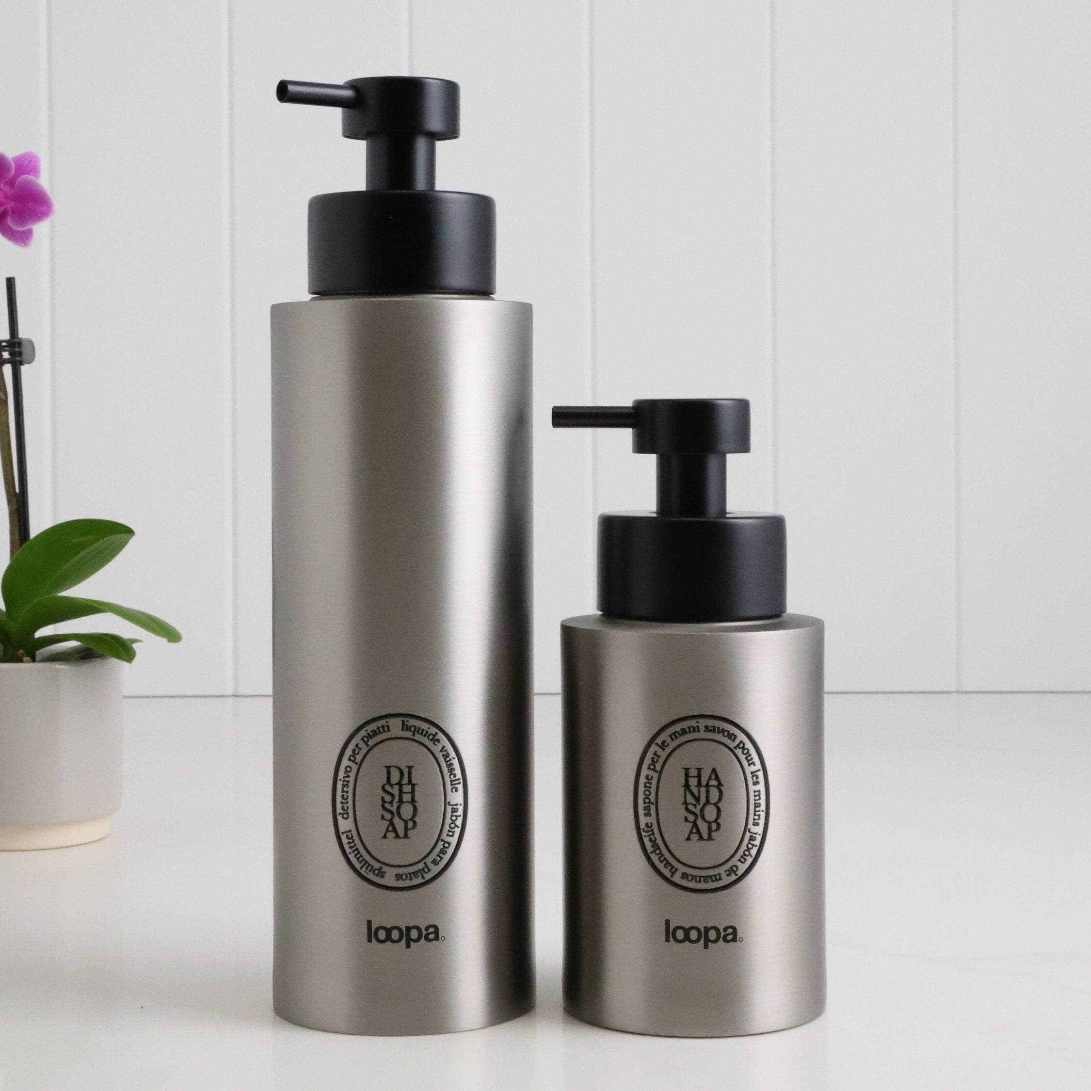 Two brushed stainless steel round bottle dispensers with black oval labels, shown together in 500ml and 250ml sizes for use across bathrooms, kitchens, or cloakroom spaces