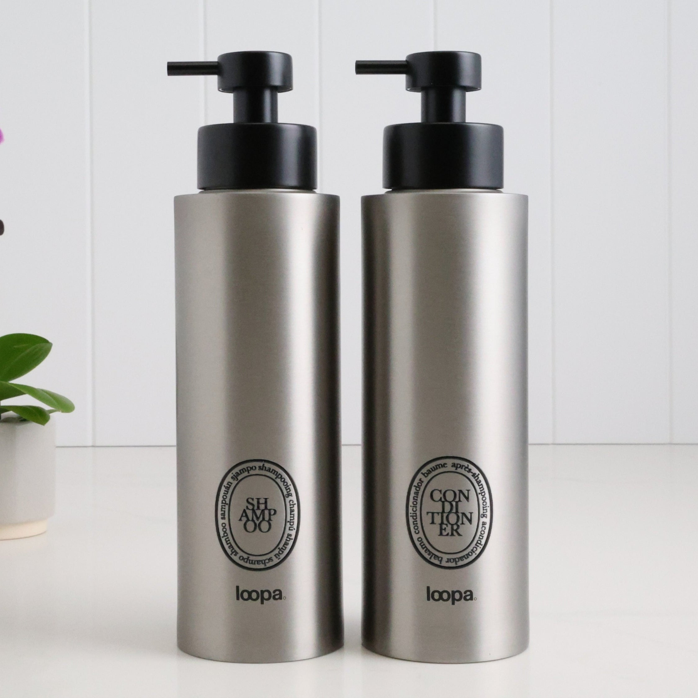Two brushed stainless steel round bottle dispensers with black oval labels, shown as matching 500ml refillable bottles suited to bathrooms, cloakrooms, or kitchen counters