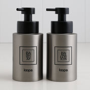 Two brushed stainless steel round bottle dispensers with black square labels, presented as matching 250ml refillable bottles for bathrooms, cloakrooms, or WC areas