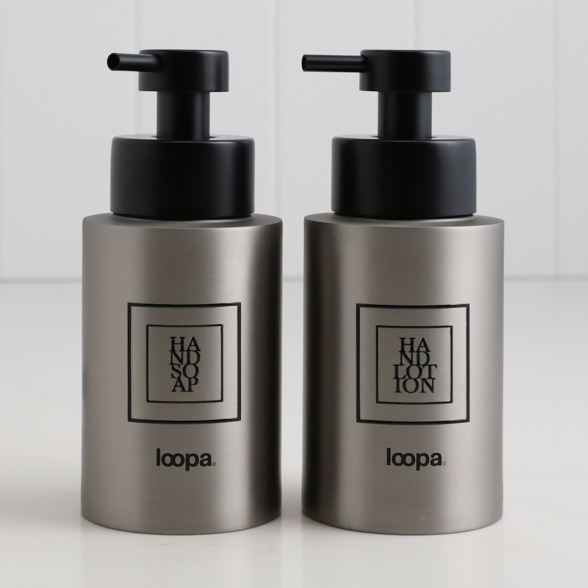 Two brushed stainless steel round bottle dispensers with black square labels, presented as matching 250ml refillable bottles for bathrooms, cloakrooms, or WC areas