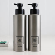 Two brushed stainless steel round bottle dispensers with black square labels, shown as matching 500ml refillable bottles for kitchen counters, bathrooms, or guest WC spaces