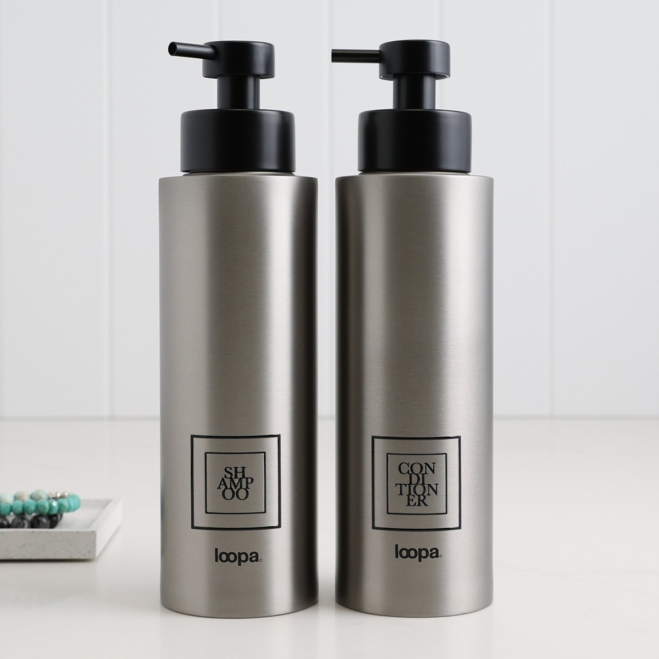 Two brushed stainless steel round bottle dispensers with black square labels, shown as matching 500ml refillable bottles for kitchen counters, bathrooms, or guest WC spaces