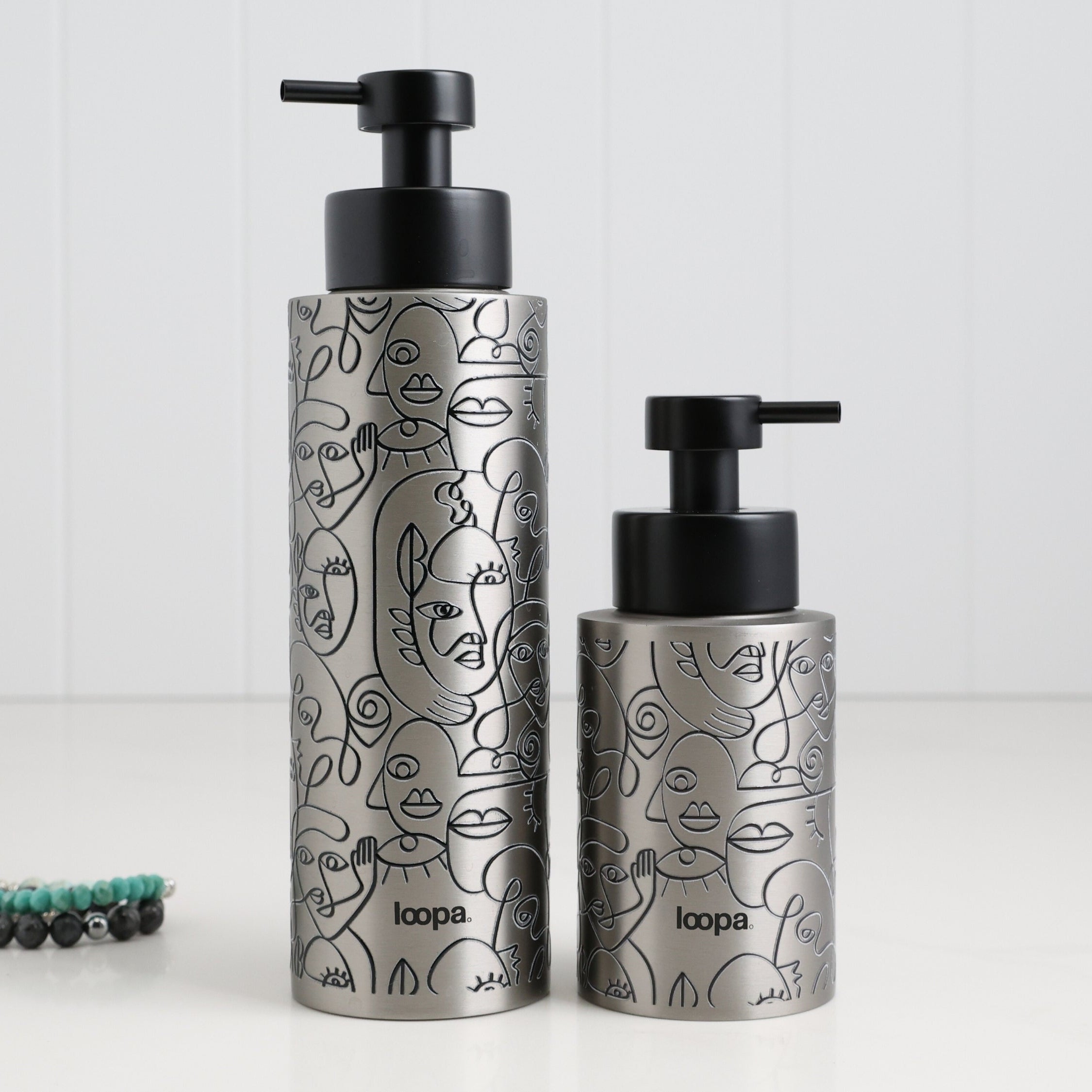 Two brushed stainless steel round bottle dispensers with black wrap labels, shown together in 250ml and 500ml sizes for coordinated sink-side use
