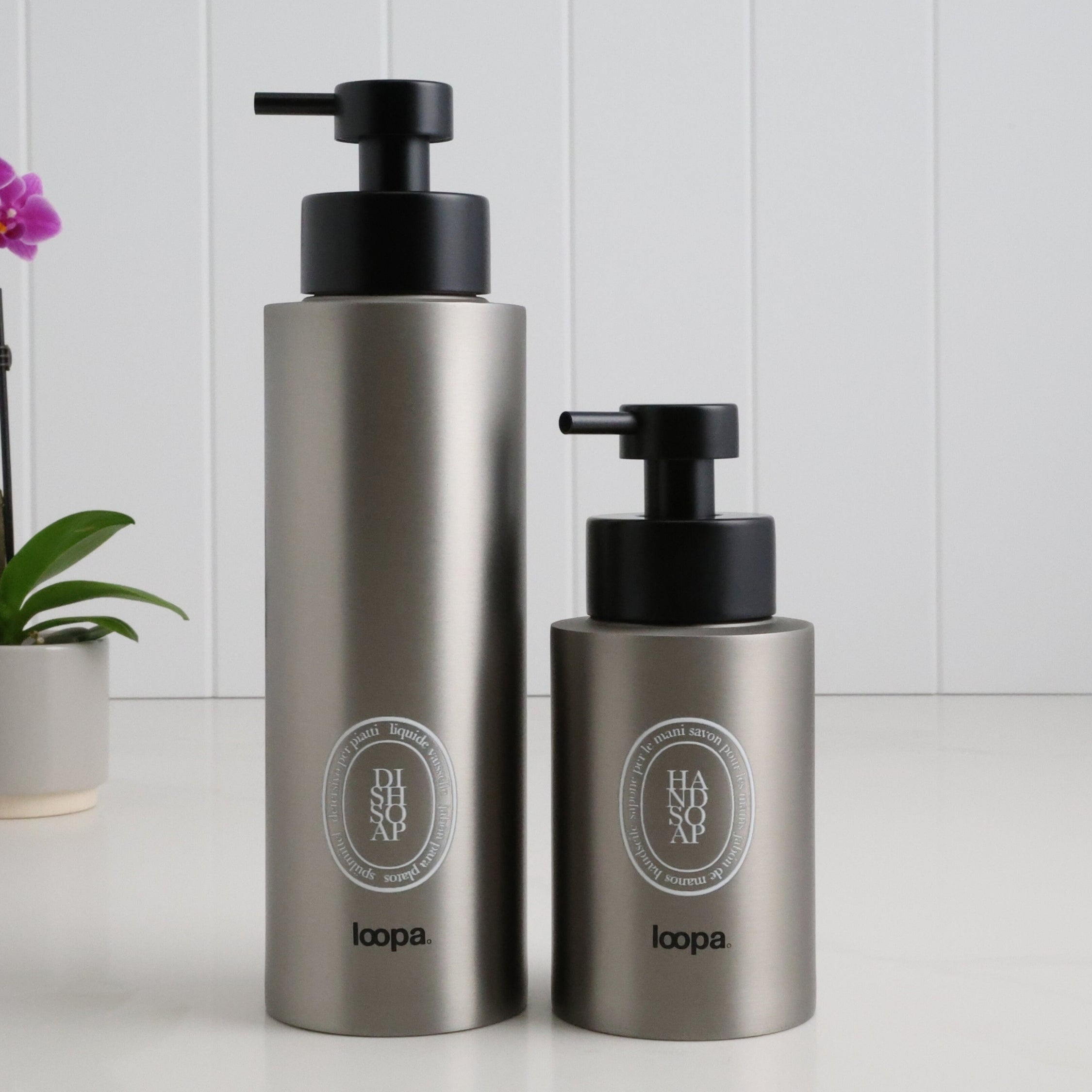 Two brushed stainless steel round bottle dispensers with white oval labels, shown together in 500ml and 250ml sizes for kitchens, bathrooms, or guest WC spaces