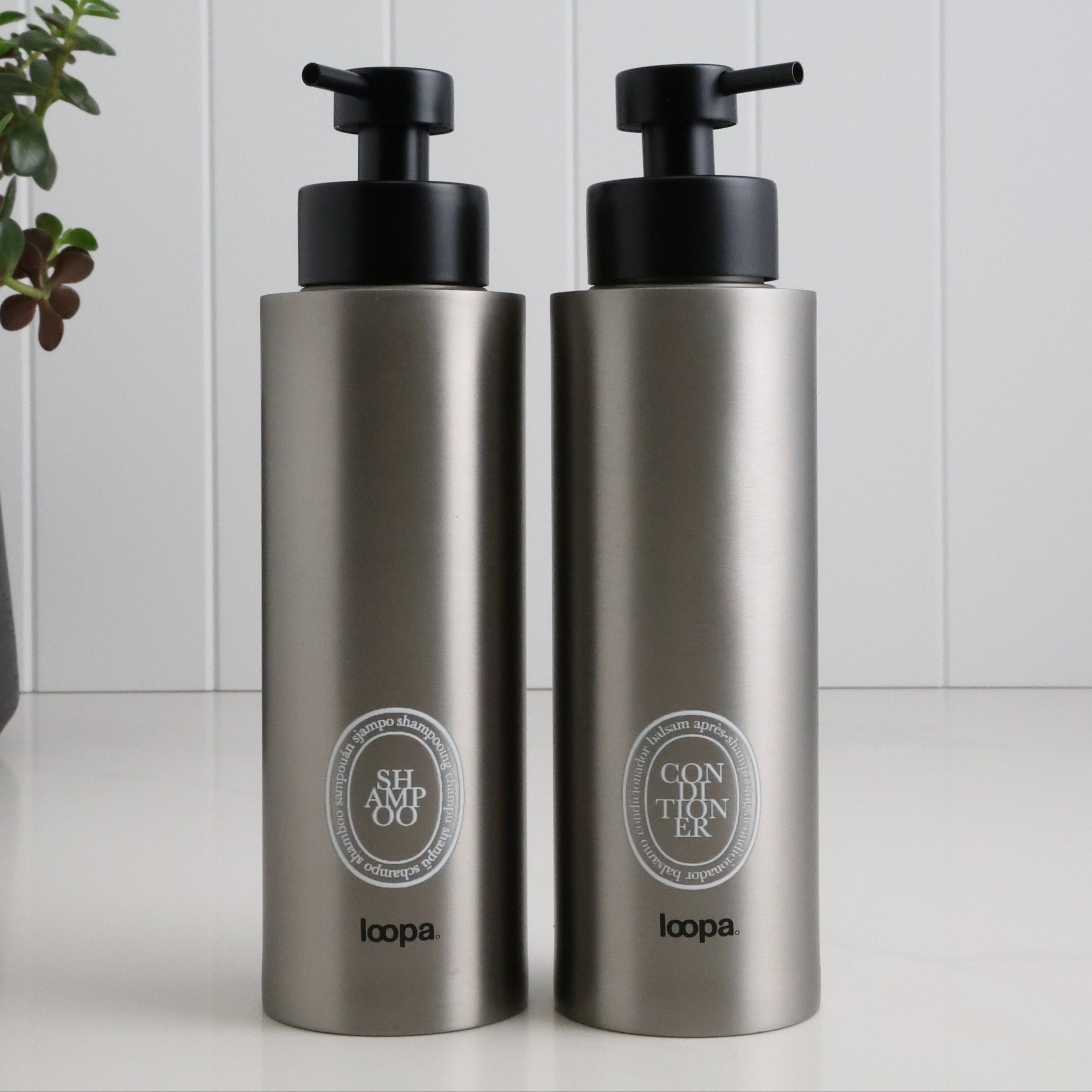 Two brushed stainless steel round bottle dispensers with white oval labels, shown as matching 500ml refillable bottles for bathrooms, shower areas, or kitchen counters