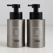Two brushed stainless steel round bottle dispensers with white square labels, shown together as matching 250ml refillable bottles for bathrooms, cloakrooms, or WC spaces