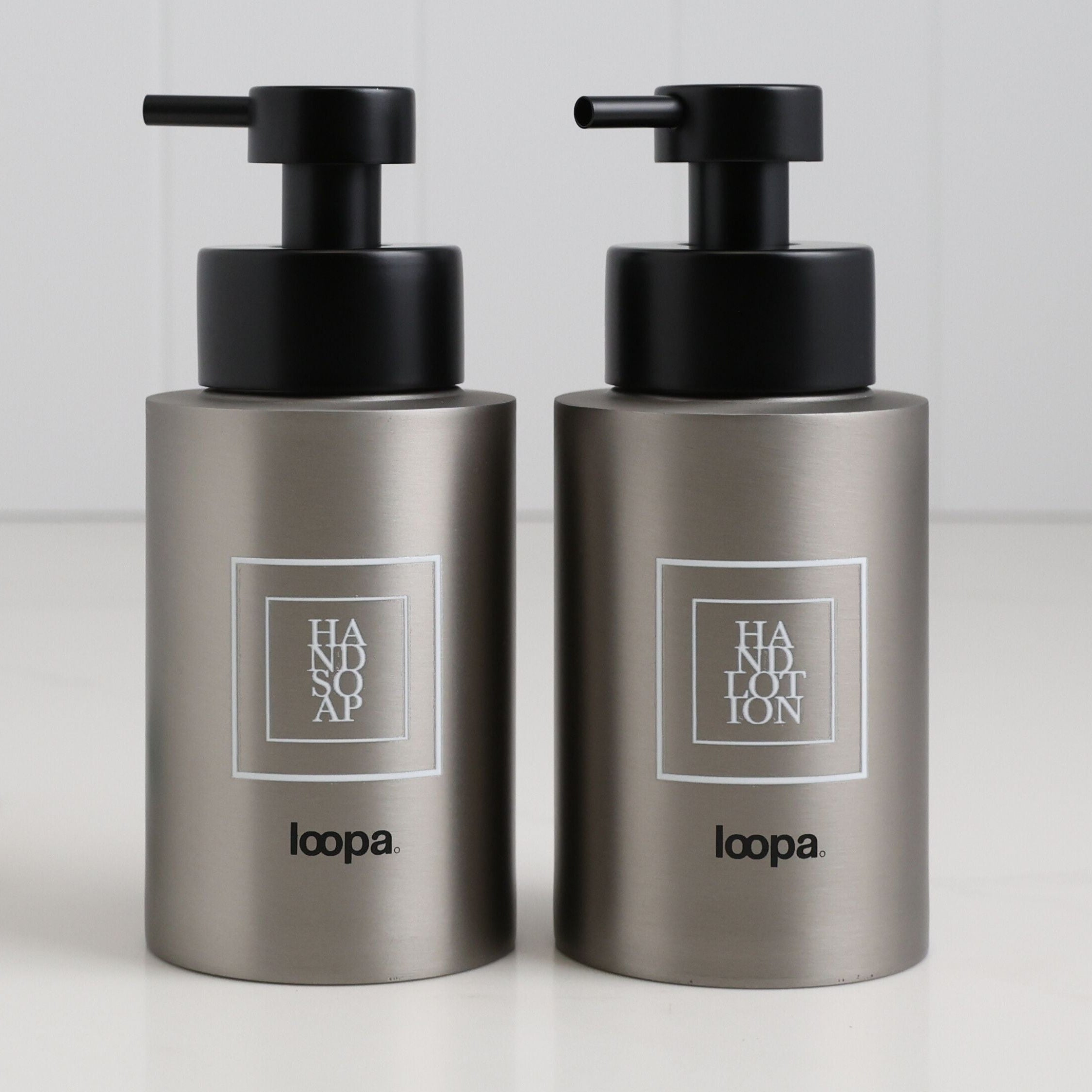 Two brushed stainless steel round bottle dispensers with white square labels, shown together as matching 250ml refillable bottles for bathrooms, cloakrooms, or WC spaces