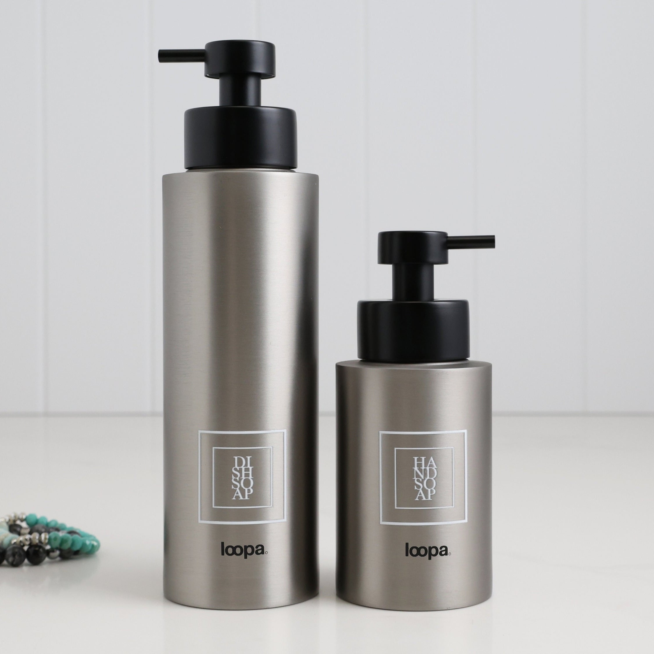 Two brushed stainless steel round bottle dispensers with white square labels, shown together in 500ml and 250ml sizes for coordinated sink-side use