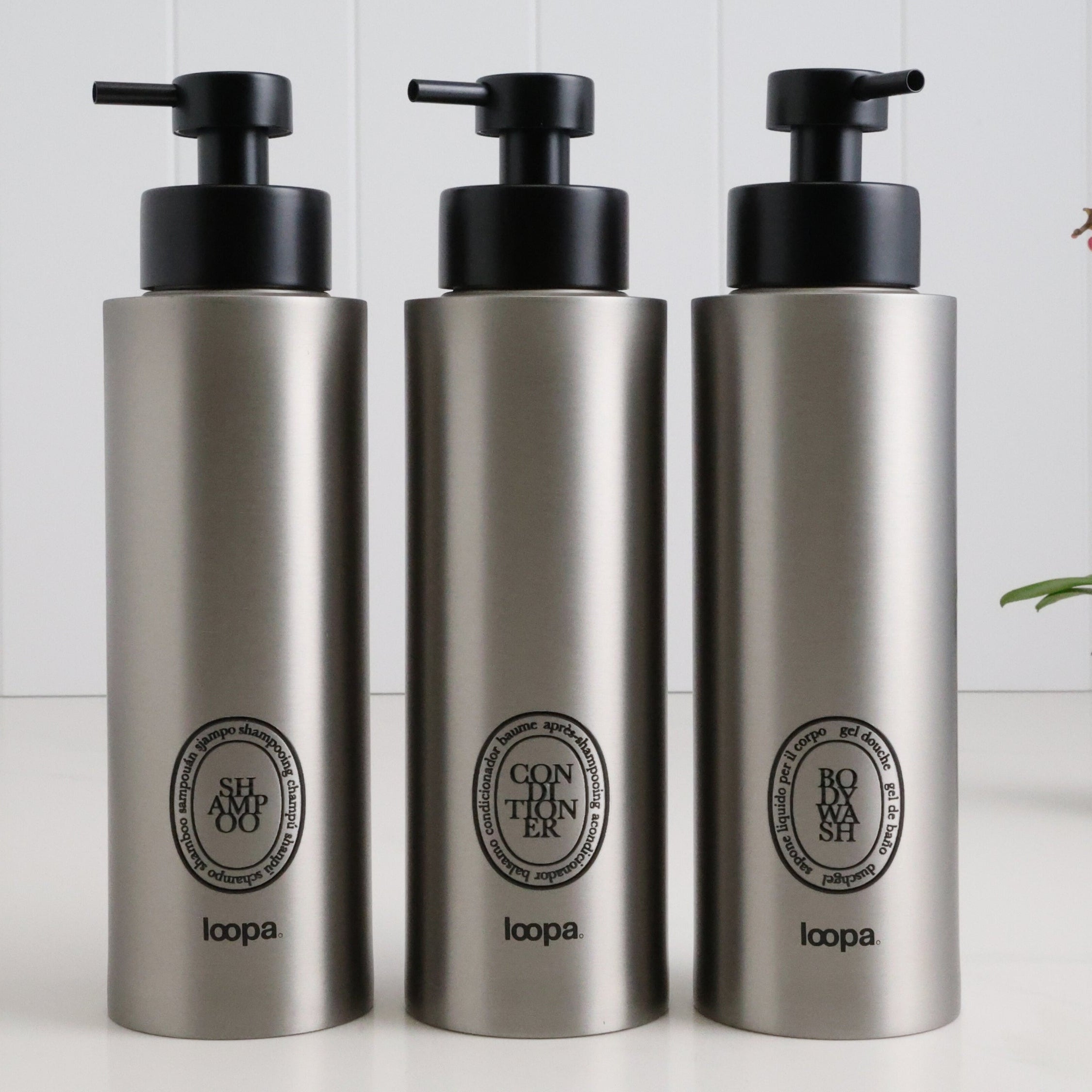 Three brushed stainless steel round bottle dispensers with black oval labels, arranged as matching 500ml refillable bottles for bathrooms, showers, or modern kitchen spaces