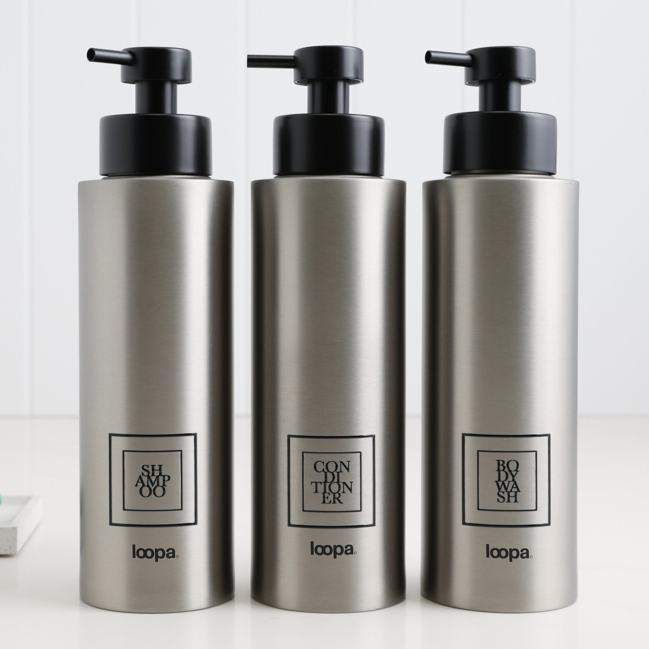 Three brushed stainless steel round bottle dispensers with black square labels, arranged as matching 500ml refillable bottles for kitchens, bathrooms, or shower spaces