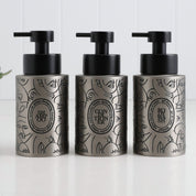 Three brushed stainless steel round bottle dispensers with boho-style black labels, shown together as matching 250ml refillable options for soap or lotion