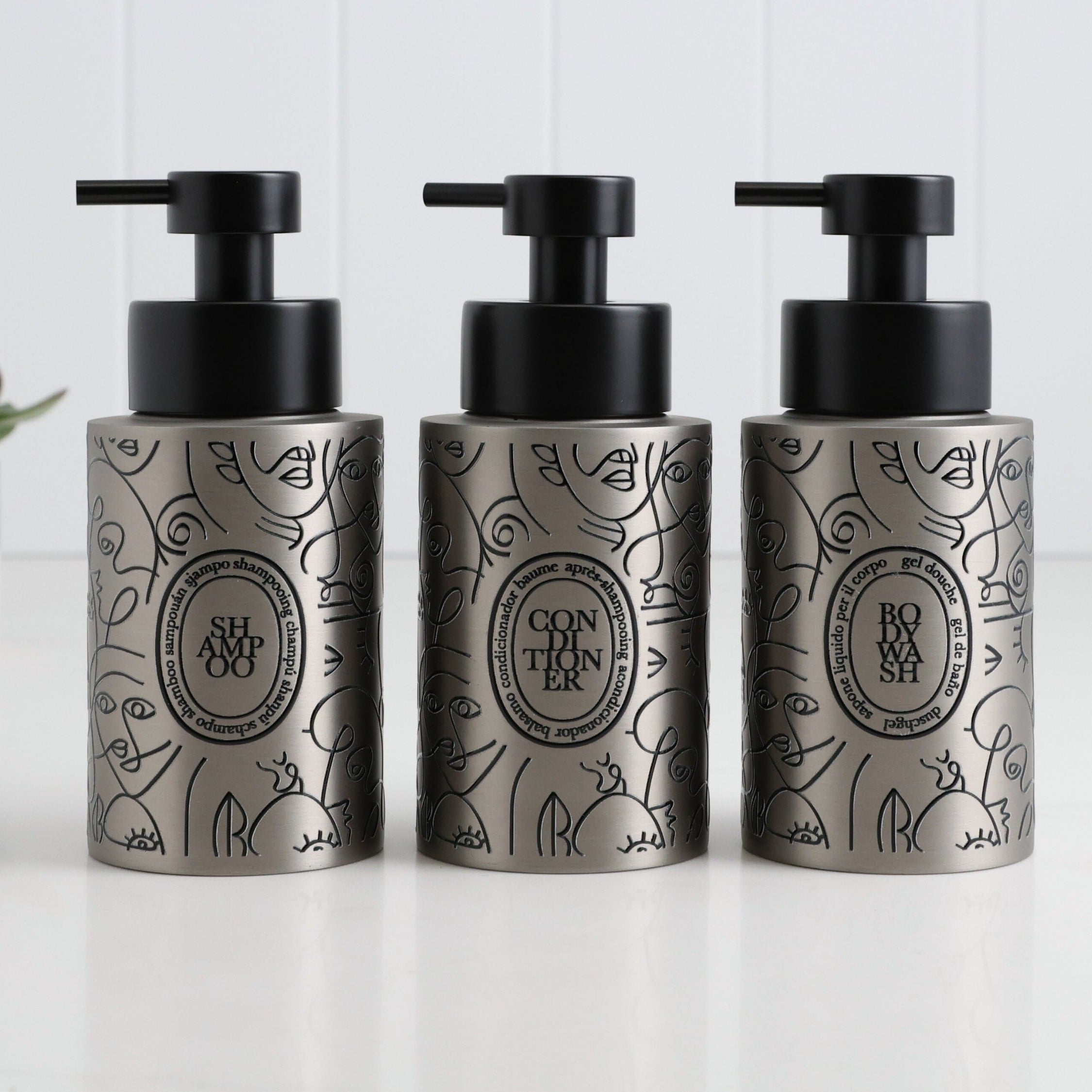 Three brushed stainless steel round bottle dispensers with boho-style black labels, shown together as matching 250ml refillable options for soap or lotion