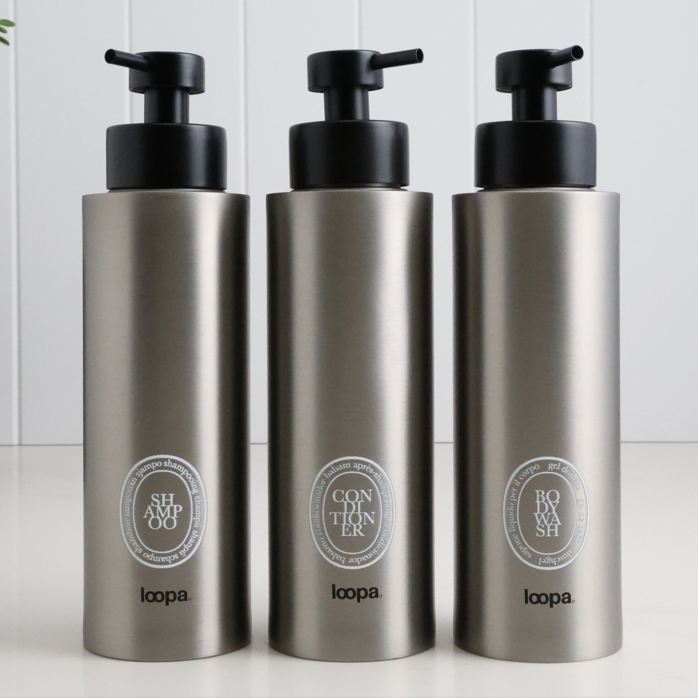Three brushed stainless steel round bottle dispensers with white oval labels, arranged as matching 500ml refillable bottles for bathrooms, showers, or modern kitchen spaces