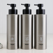Three brushed stainless steel round bottle dispensers with white square labels, shown as matching 500ml refillable bottles arranged for modern sink areas
