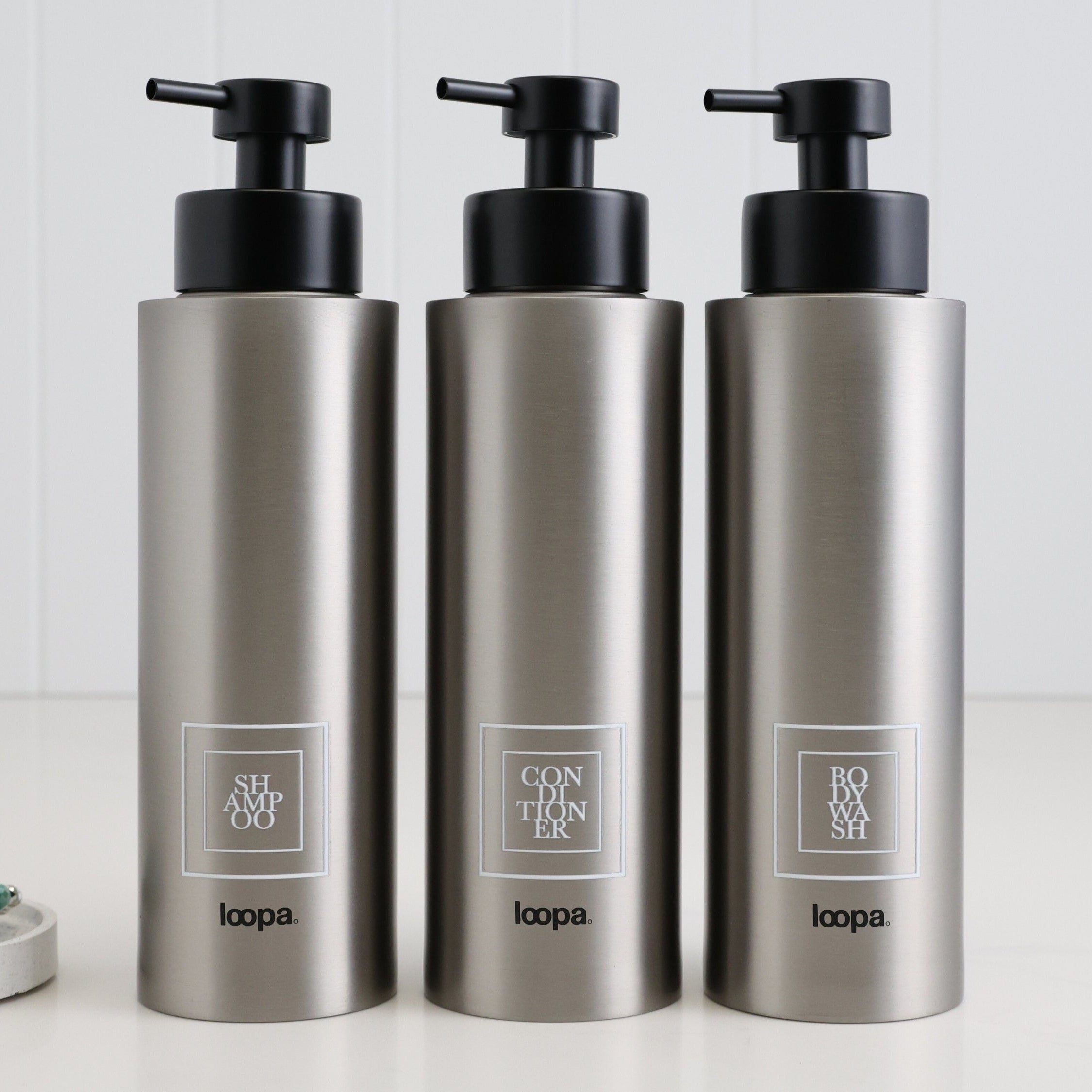 Three brushed stainless steel round bottle dispensers with white square labels, shown as matching 500ml refillable bottles arranged for modern sink areas