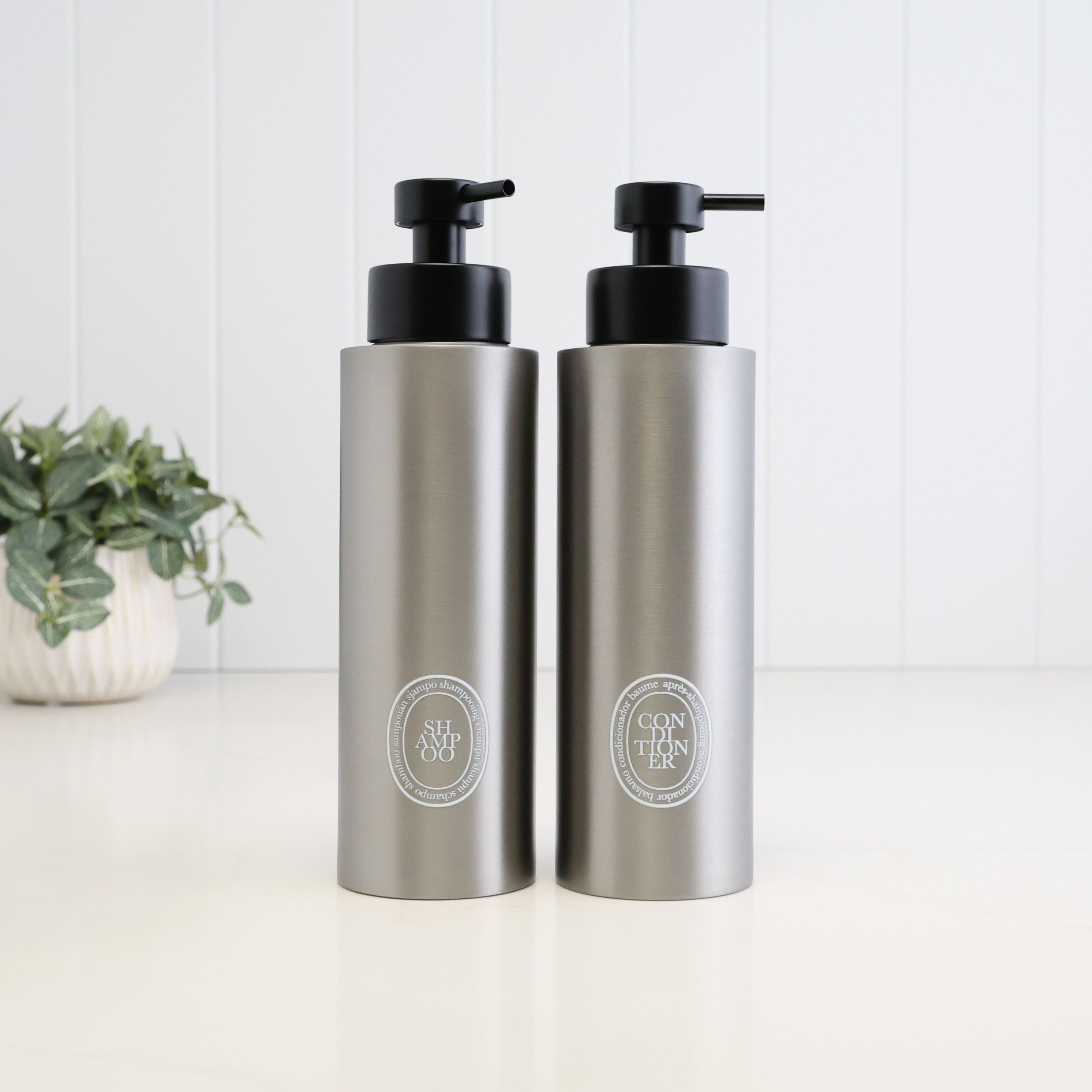 Pair of 500ml brushed 304 stainless steel shampoo and conditioner dispensers with white oval UV-DTF labels and satin black liquid pumps, designed as a refillable bathroom and shower bottle set.