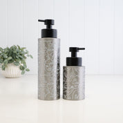 Two brushed silver metal pump bottles with black pumps, decorated with white line art illustrations, standing on a white surface against a white paneled background with a small potted plant on the left.
