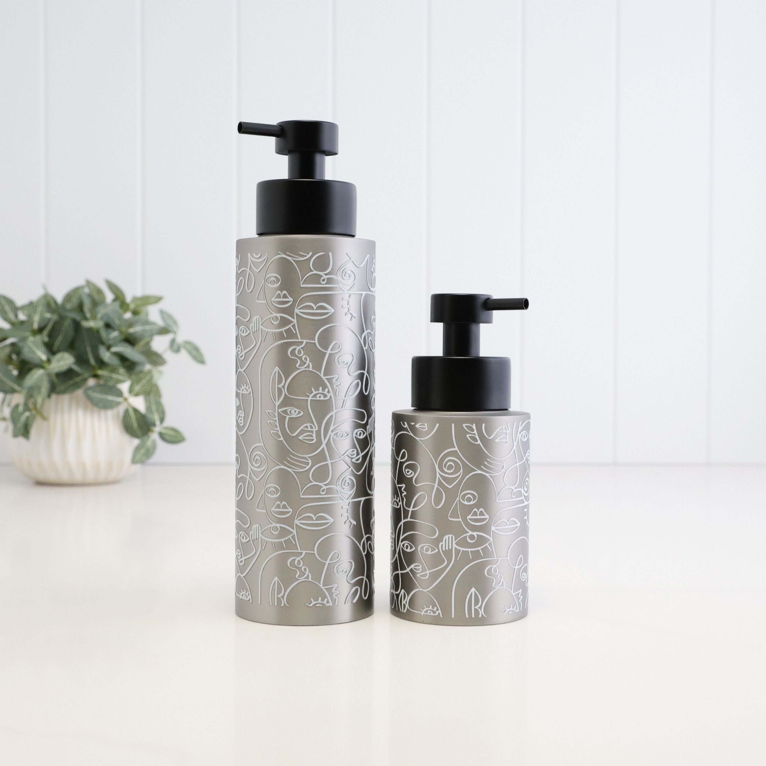 Two brushed silver metal pump bottles with black pumps, decorated with white line art illustrations, standing on a white surface against a white paneled background with a small potted plant on the left.