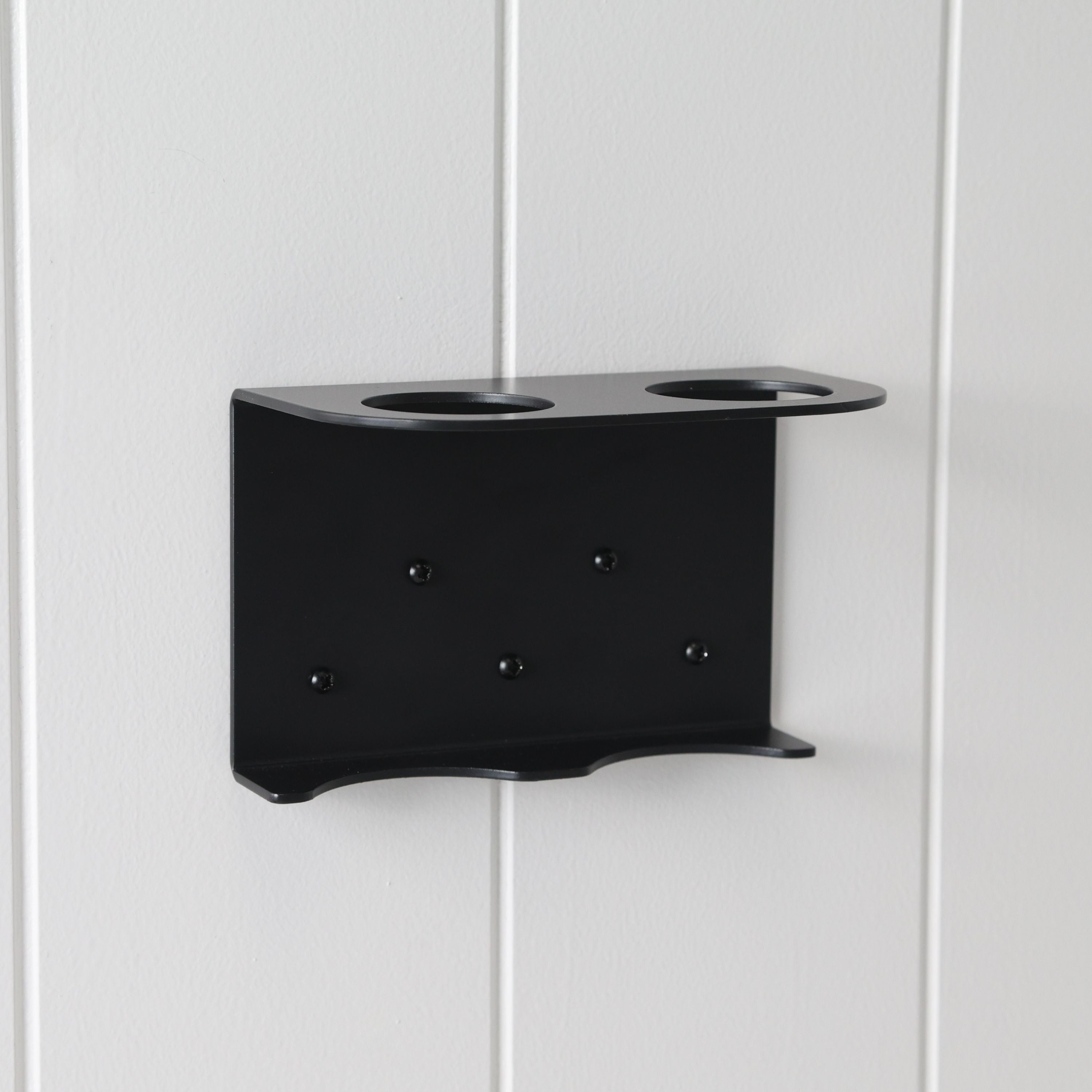 Loopa double wall-mounted soap dispenser bracket in black powder-coated aluminium, securely installed on a white panelled wall. Featuring two circular bottle openings and a supporting base, this minimalist metal holder accommodates two Loopa 250ml or 500ml refillable dispenser bottles. Ideal for organising hand soap and lotion in bathrooms or shampoo and conditioner in showers.