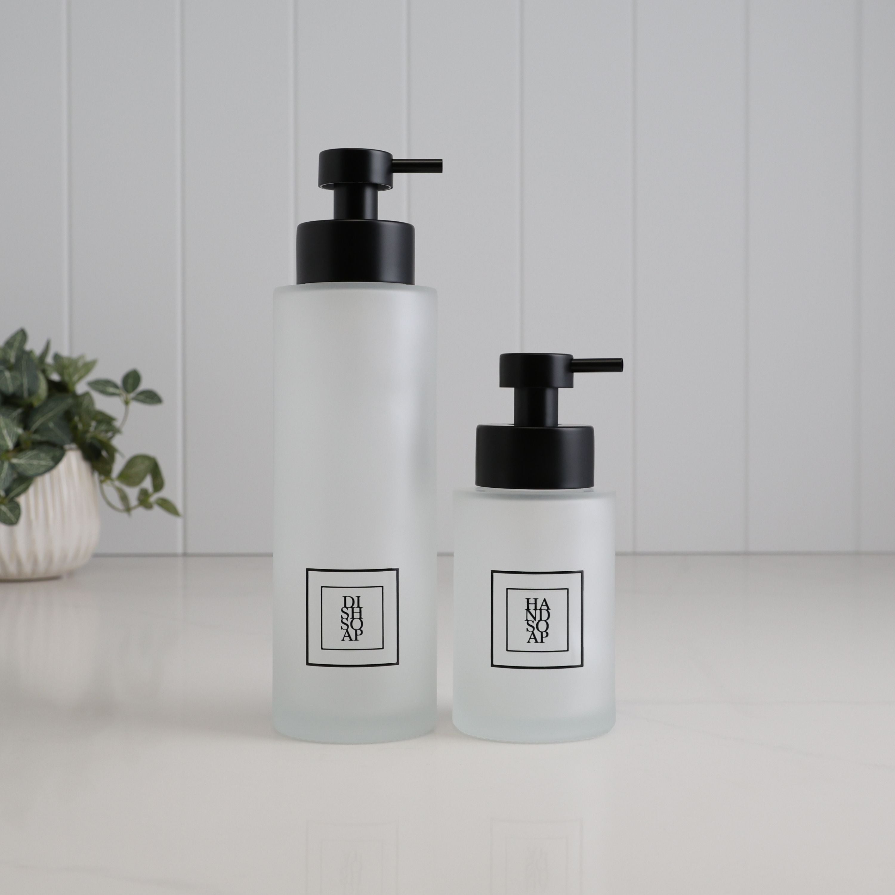 Matching frosted glass dispenser set with tall 500ml dish soap bottle and 250ml hand soap bottle, finished with black square labels and satin black stainless steel pumps for a coordinated kitchen sink setup.