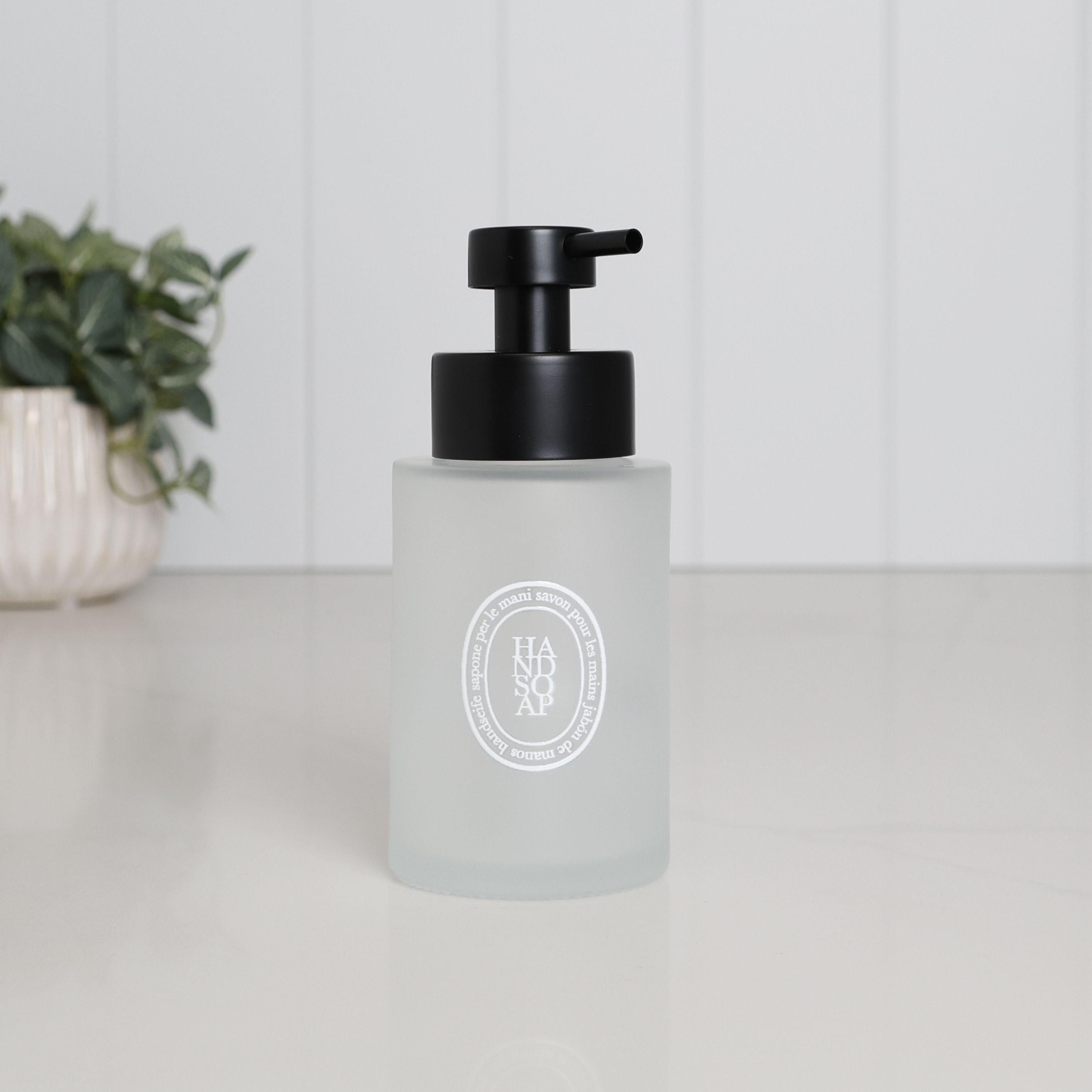 250ml frosted glass hand soap dispenser with white oval label and satin black stainless steel liquid pump, styled on a clean bathroom countertop.