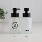 Two frosted glass pump bottles shown side by side on a white surface against a white paneled wall. The left bottle is labeled Front and shows a black pump and a circular black logo; the right bottle is labeled Back and shows the word 'loopa' in black.