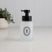 A frosted glass hand soap dispenser with a black pump and a circular black logo on the front, set on a white surface with a blurred potted plant in the background.