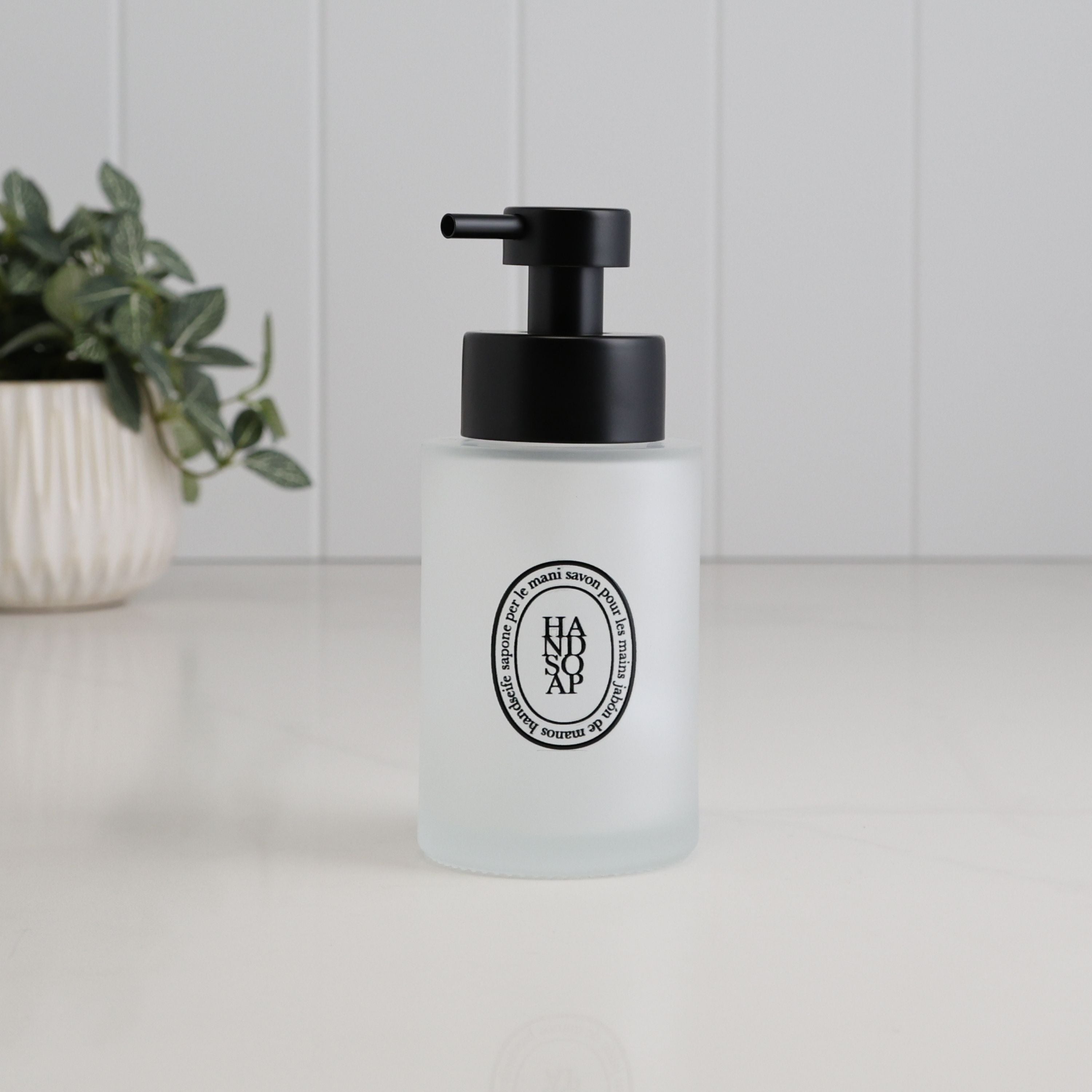 A frosted glass hand soap dispenser with a black pump and a circular black logo on the front, set on a white surface with a blurred potted plant in the background.