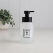 Frosted glass hand soap dispenser with black square label and satin black stainless steel pump, styled on a bathroom countertop with a plant in the background.