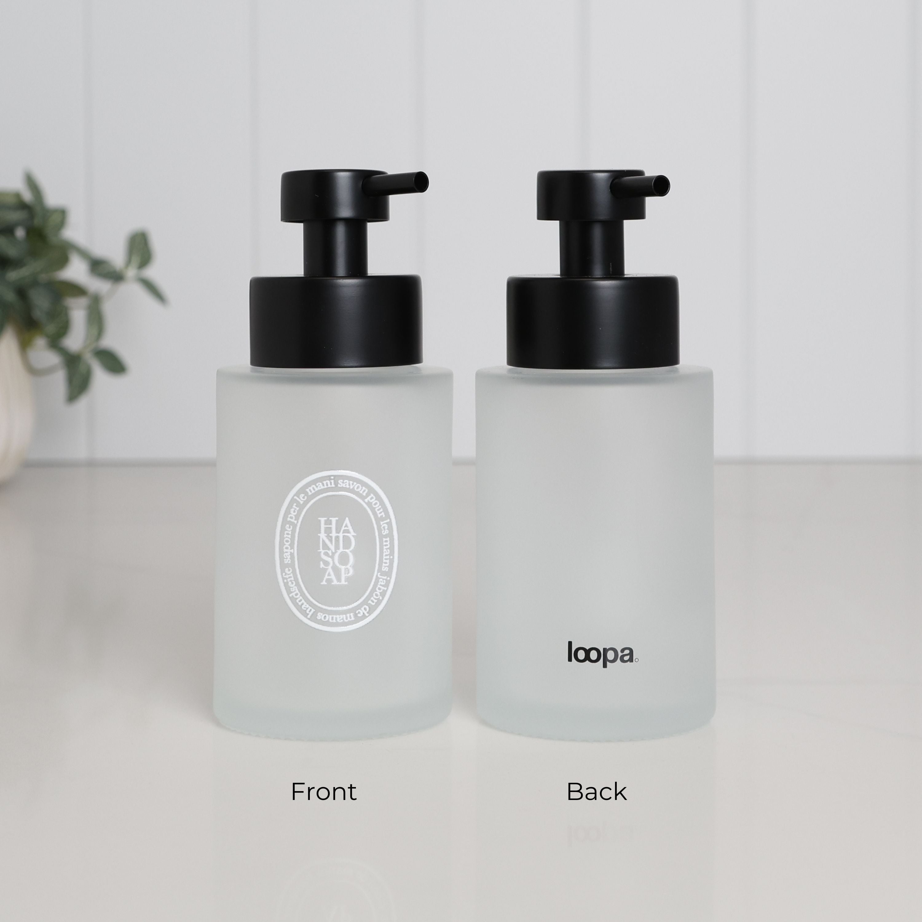 Front and rear view of a frosted glass hand soap dispenser with white oval label, Loopa logo and satin black stainless steel liquid pump.