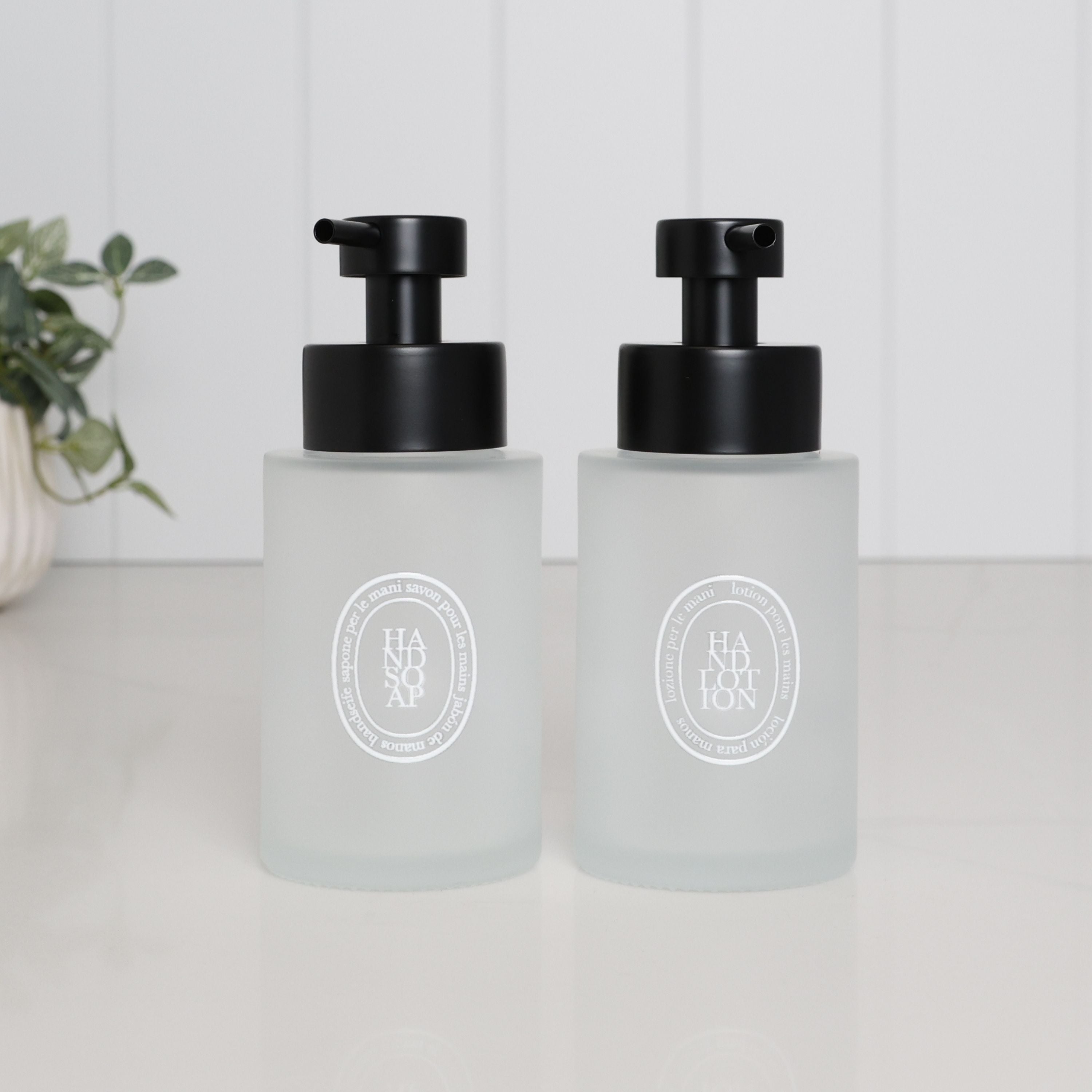 Set of two 250ml frosted glass soap dispensers for hand soap and hand lotion, with white oval labels and satin black stainless steel pumps.