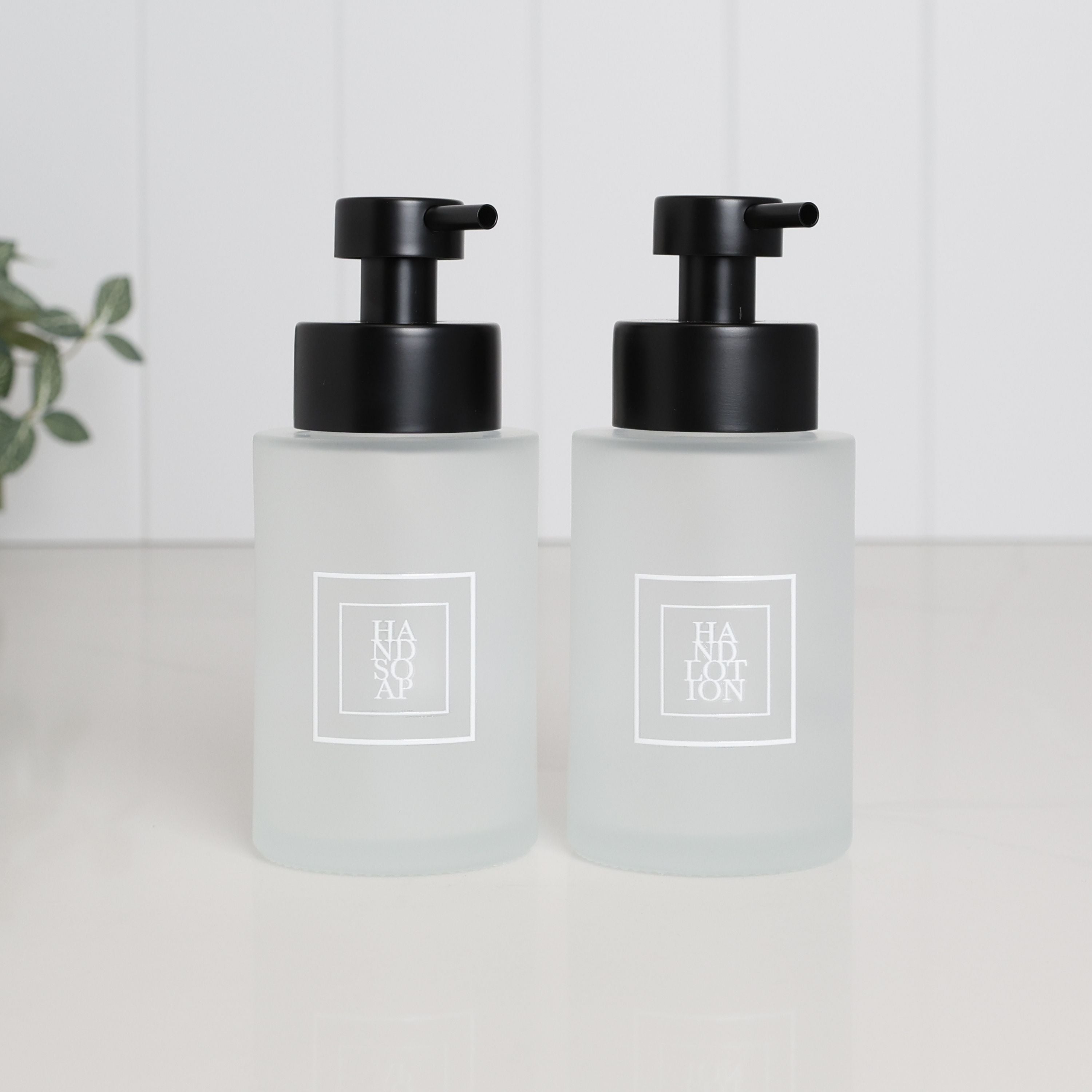 Pair of 250ml frosted glass soap dispensers with white square Hand Soap and Hand Lotion labels and satin black pumps, designed as matching refillable bathroom bottles for a sink or vanity.
