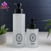 Frosted glass round bottle dispenser with a black oval label, designed for refillable soap or lotion in modern kitchen or bathroom spaces