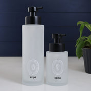 Frosted glass round bottle dispenser with a white oval label, designed for refillable soap or lotion in modern kitchen or bathroom spaces