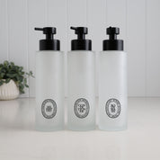 Set of three 500ml frosted glass dispensers for shampoo, conditioner and body wash, featuring black oval labels and satin black pumps for a coordinated refillable shower bottle set.