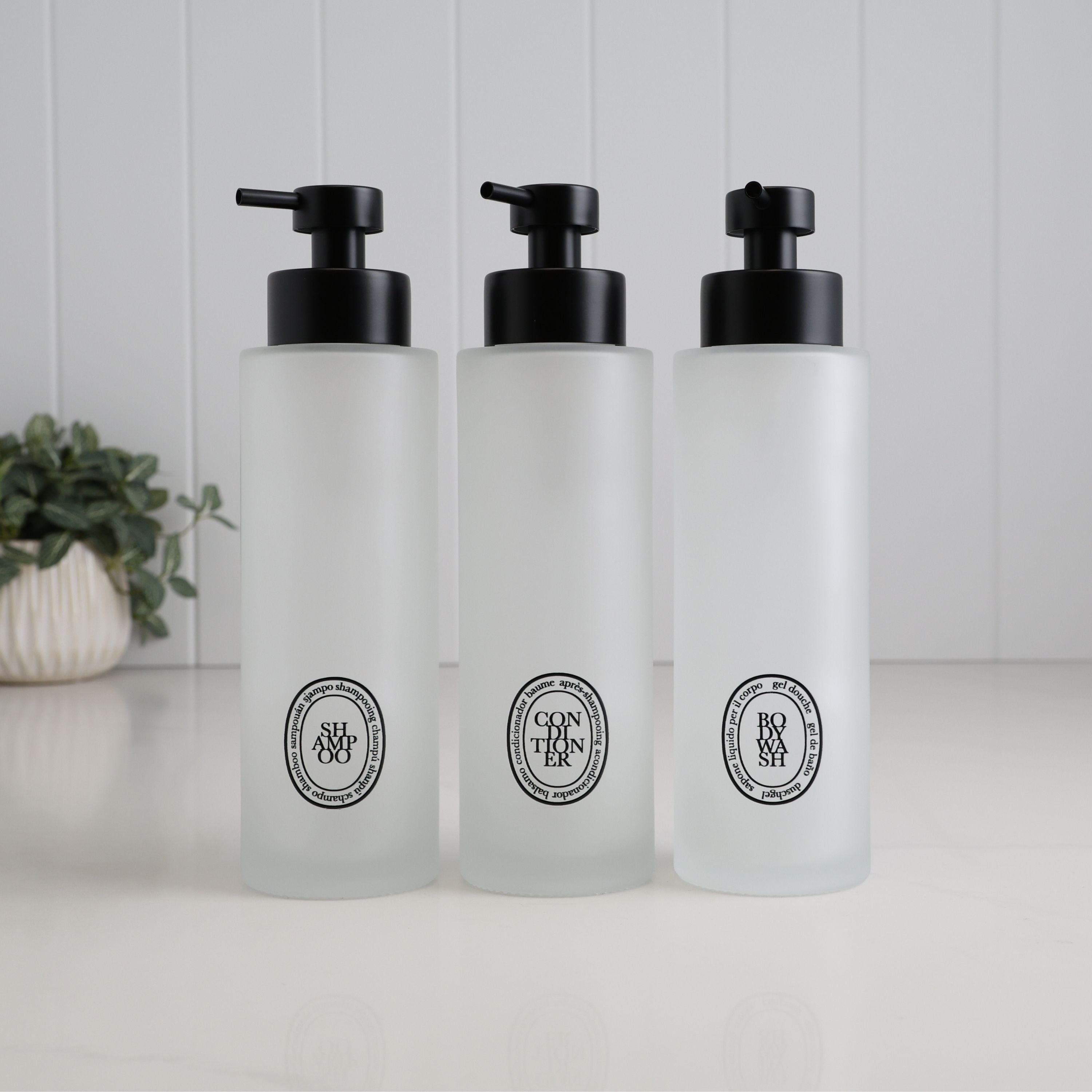 Set of three 500ml frosted glass dispensers for shampoo, conditioner and body wash, featuring black oval labels and satin black pumps for a coordinated refillable shower bottle set.