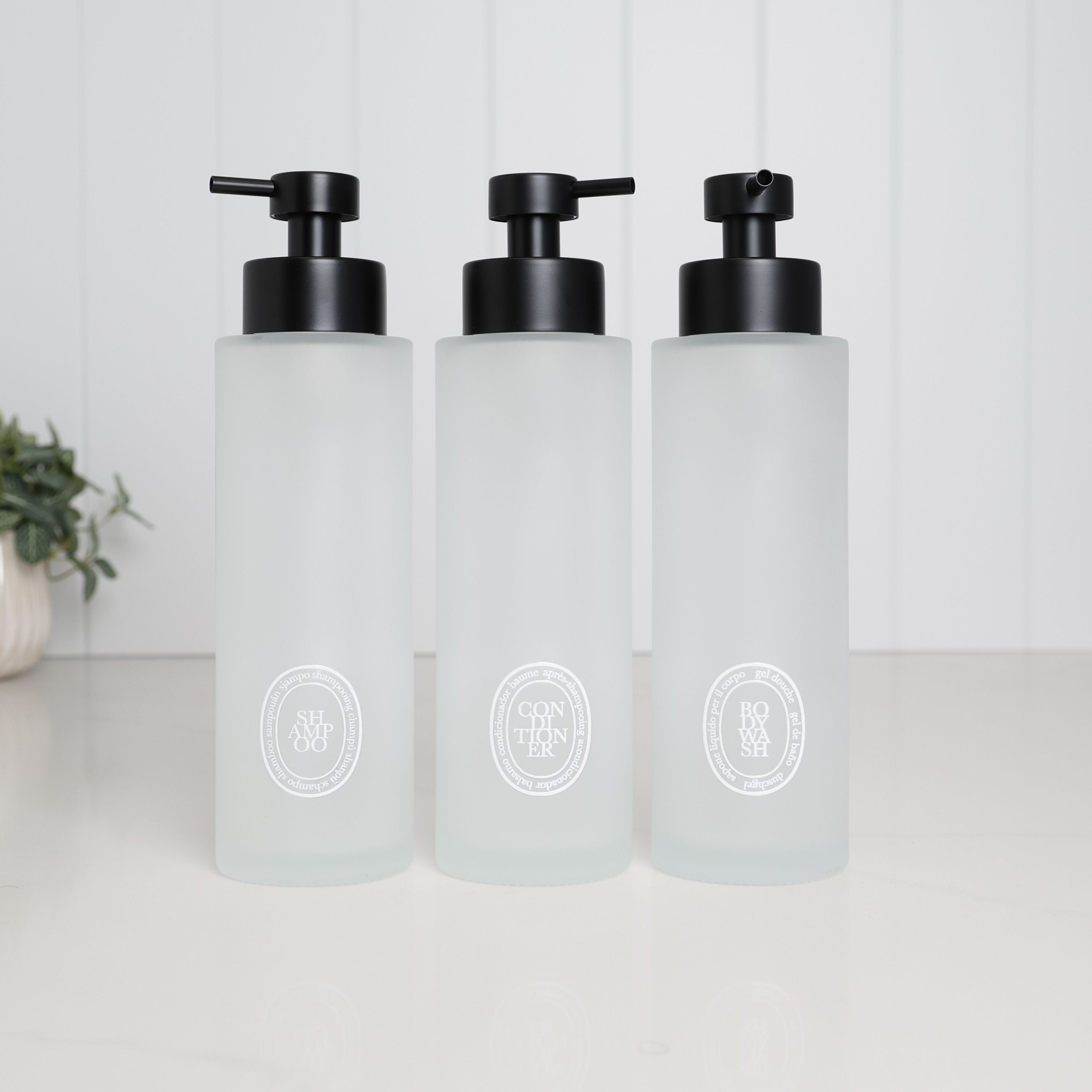 Set of three 500ml frosted glass dispensers for shampoo, conditioner and body wash, with white oval labels and satin black stainless steel pumps.