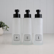 Set of three 500ml frosted glass soap dispensers for shampoo, conditioner and body wash, featuring black square labels and satin black stainless steel pumps for a coordinated bathroom or shower.