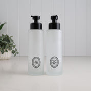 Pair of 500ml frosted glass shampoo and conditioner dispensers with black oval labels and satin black pumps, creating a matching refillable bottle set for the bathroom or shower.