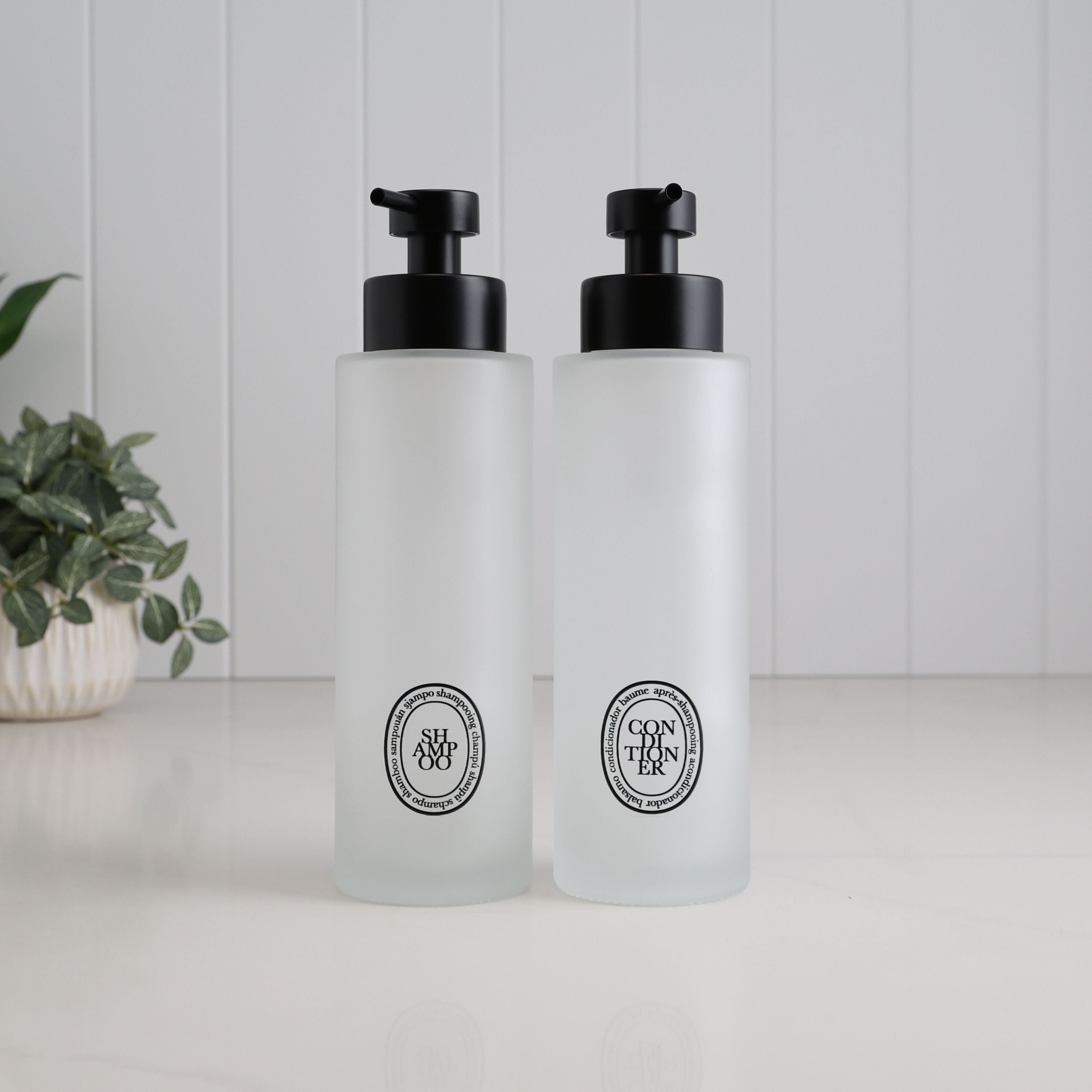 Pair of 500ml frosted glass shampoo and conditioner dispensers with black oval labels and satin black pumps, creating a matching refillable bottle set for the bathroom or shower.