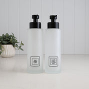 Set of two 500ml frosted glass shampoo and conditioner dispensers with black square labels and satin black stainless steel pumps, designed for a modern bathroom or shower.