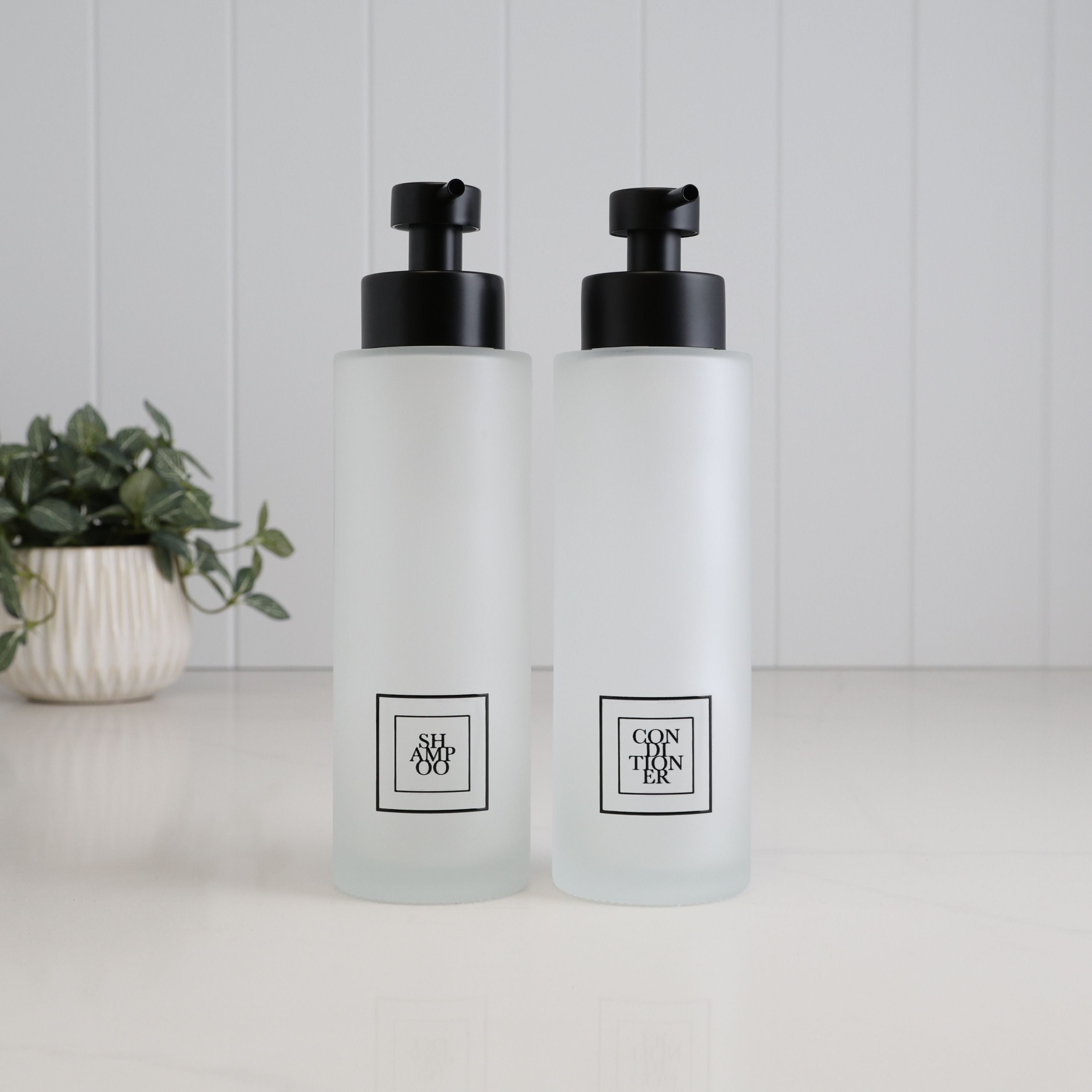 Set of two 500ml frosted glass shampoo and conditioner dispensers with black square labels and satin black stainless steel pumps, designed for a modern bathroom or shower.