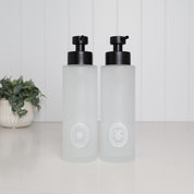 Set of two 500ml frosted glass shampoo and conditioner dispensers with white oval labels and satin black stainless steel pumps.