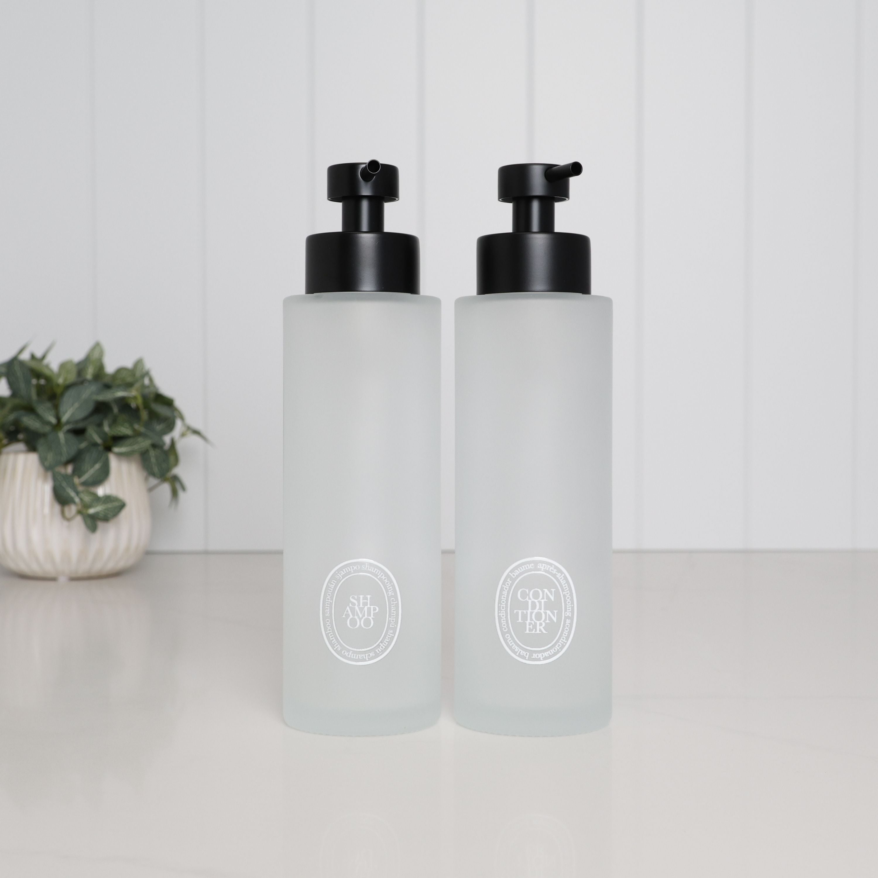 Set of two 500ml frosted glass shampoo and conditioner dispensers with white oval labels and satin black stainless steel pumps.