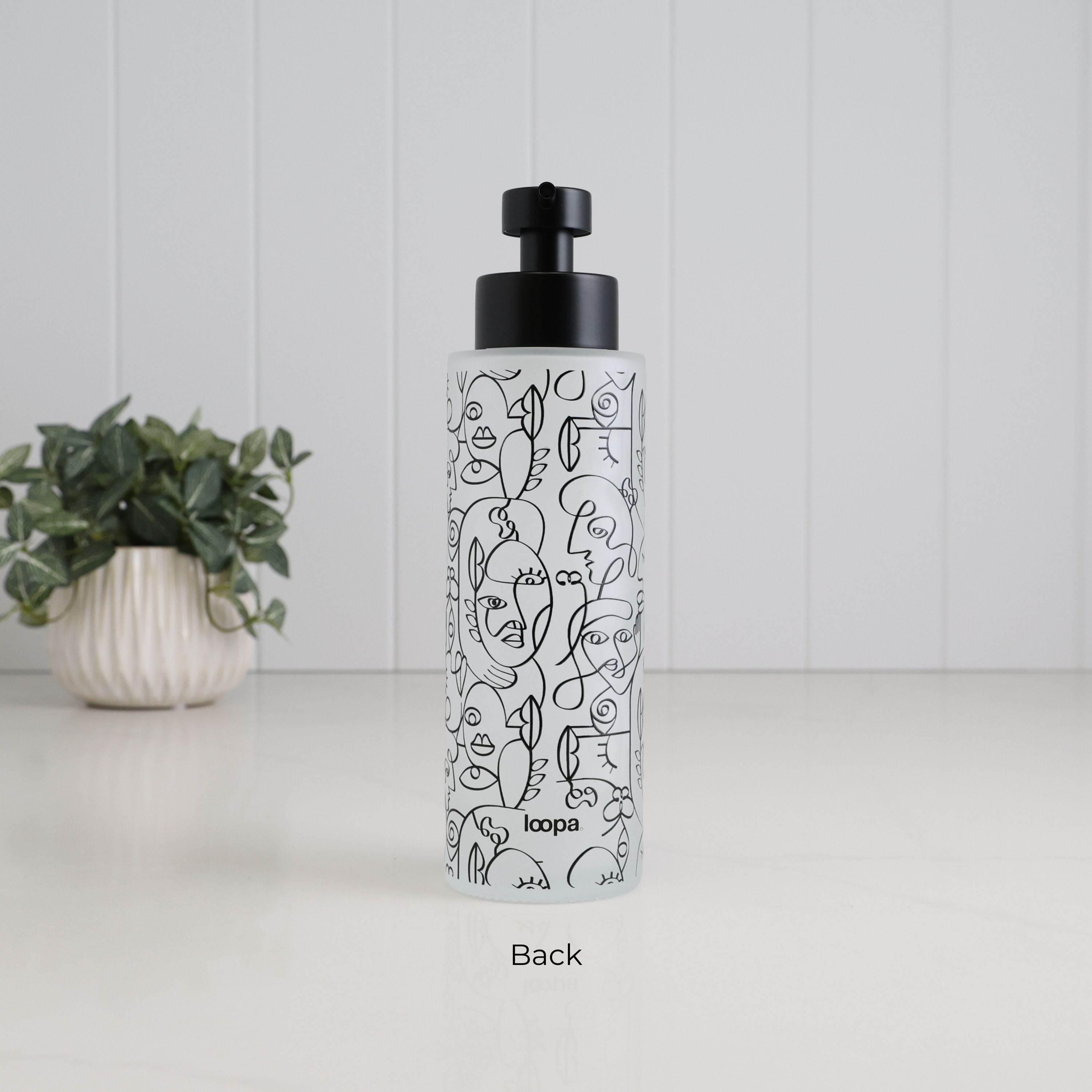 Back view of a tall frosted glass soap dispenser with black abstract face line art, Loopa branding and satin black pump, showing the decorative finish of the refillable 500ml bathroom bottle.