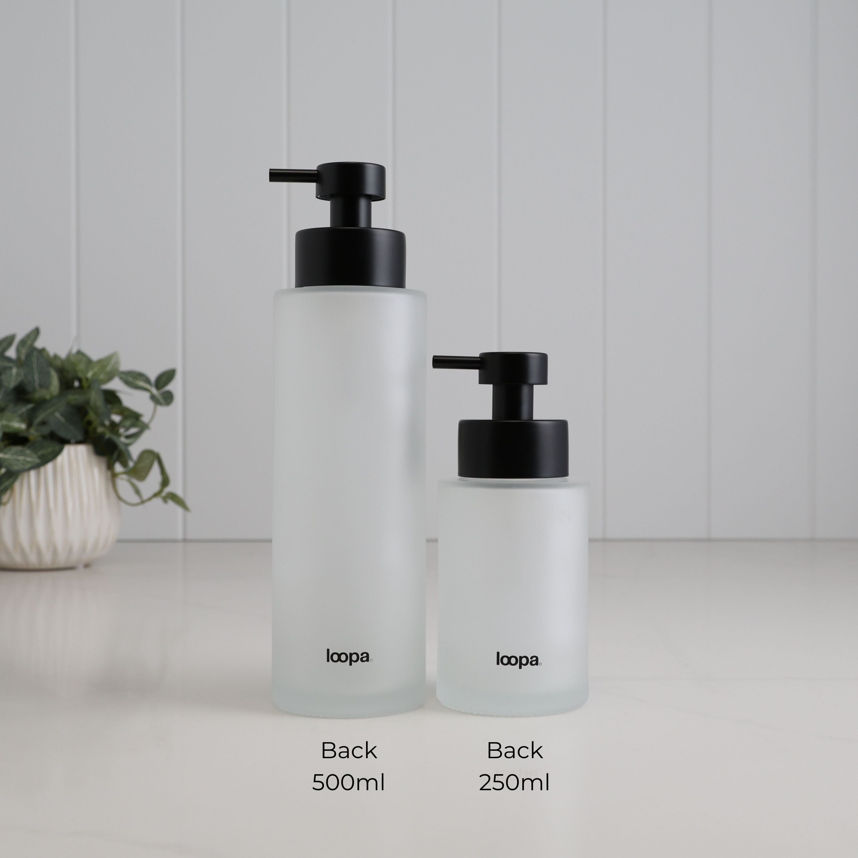 Back view of 500ml and 250ml frosted glass soap dispensers shown side by side for size comparison, with Loopa branding and satin black stainless steel pumps.