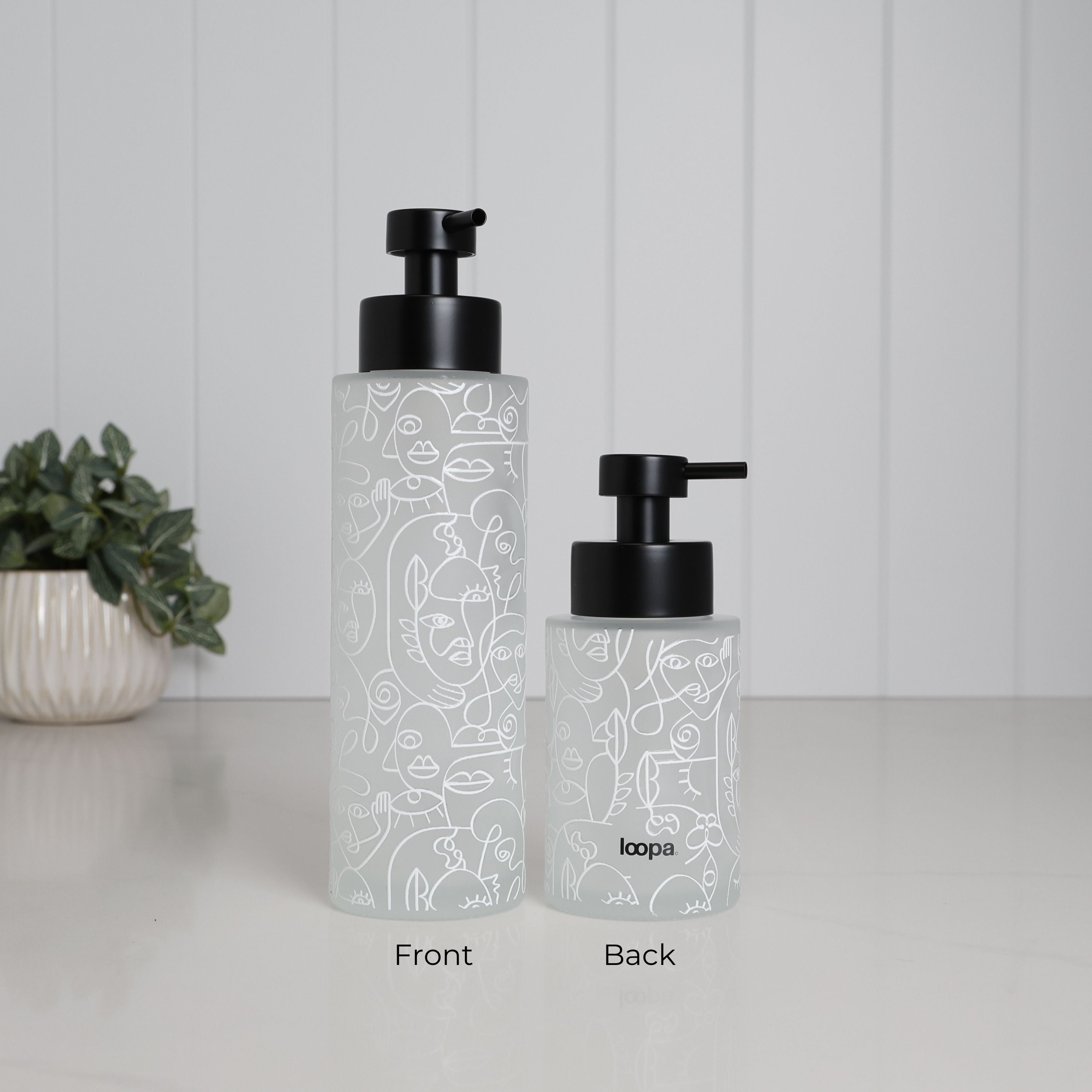 Front and back view of frosted glass soap dispenser set with abstract face line art wrap, 500ml tall bottle and 250ml small bottle with matte black stainless steel pumps, refillable bathroom accessories for hand soap lotion or shampoo, modern decorative glass dispensers for minimalist sink or vanity styling