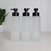 Set of three 500ml frosted glass soap dispensers with white shampoo, conditioner and body wash labels and satin black pumps, designed as refillable bathroom bottles for a modern shower or countertop.